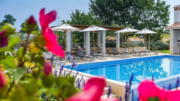 Ein Pool mit Liegestühlen und Sonnenschirmen, umgeben von Grünflächen und einer Pergola.