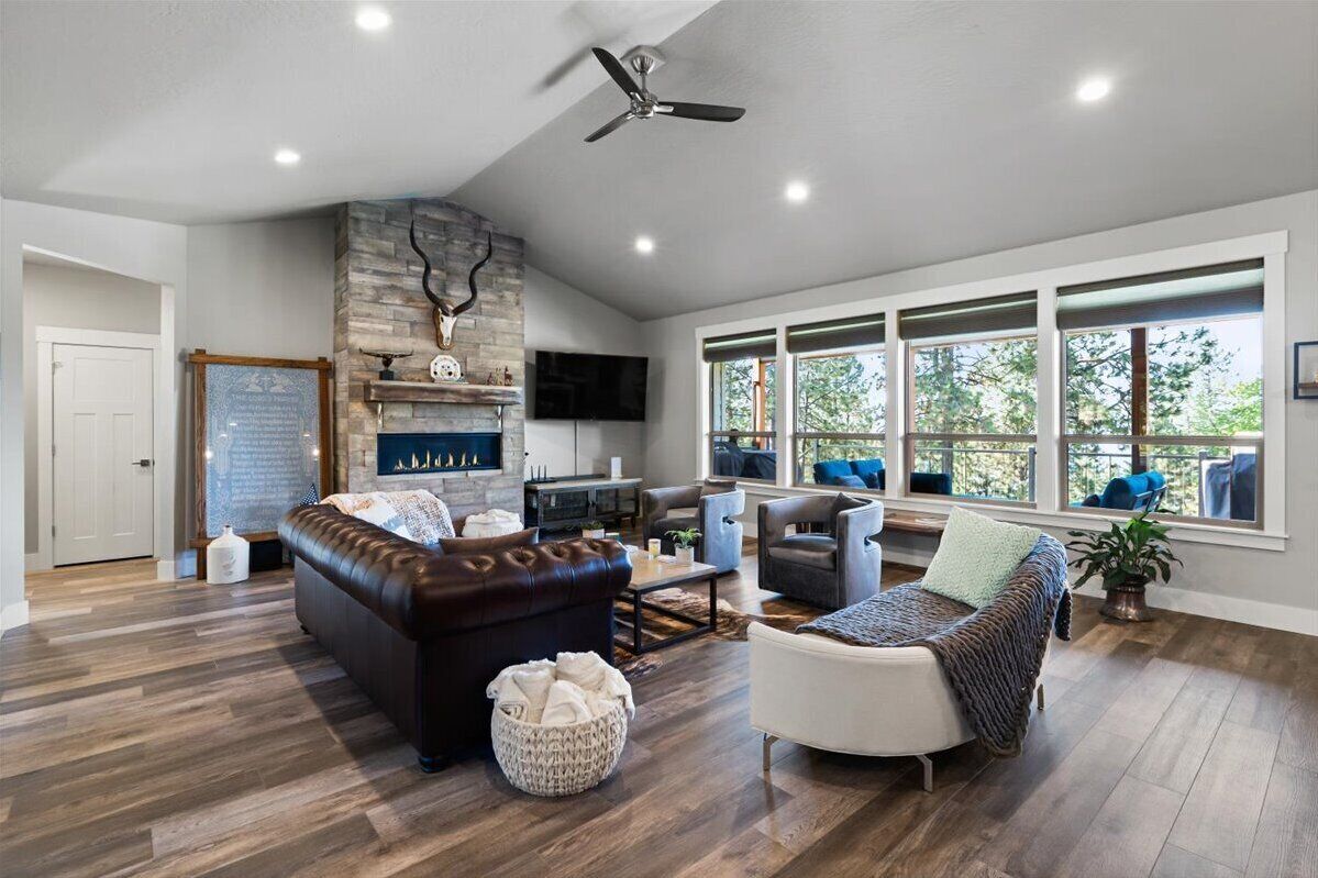 A spacious living room with a stone fireplace, a ceiling fan, and large windows offering a view of the outdoors.