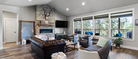 A spacious living room with a stone fireplace, a ceiling fan, and large windows offering a view of the outdoors.