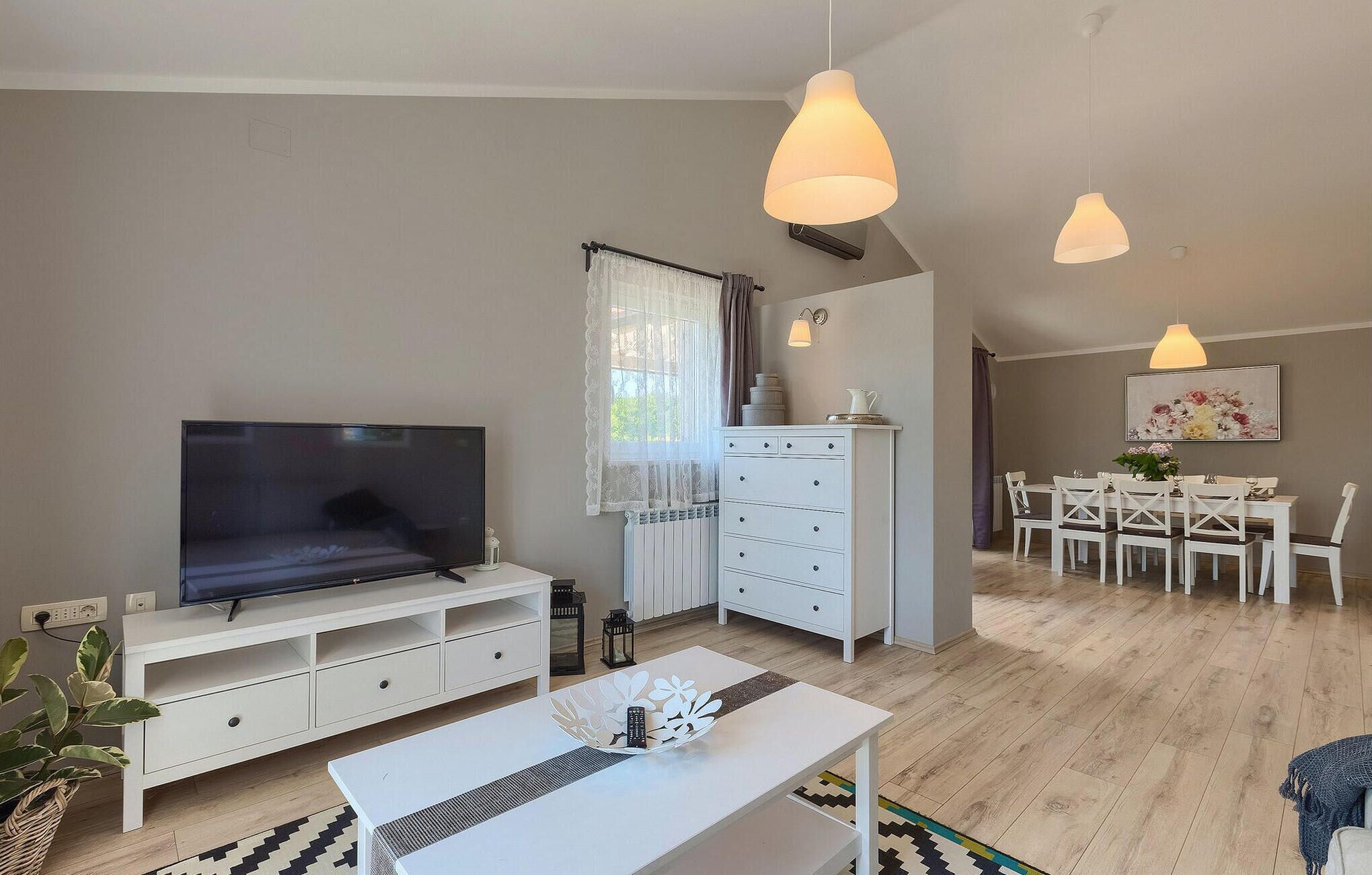 Een moderne woonkamer met een flatscreentelevisie, witte meubels, een eethoek met een tafel en stoelen, en hanglampen.