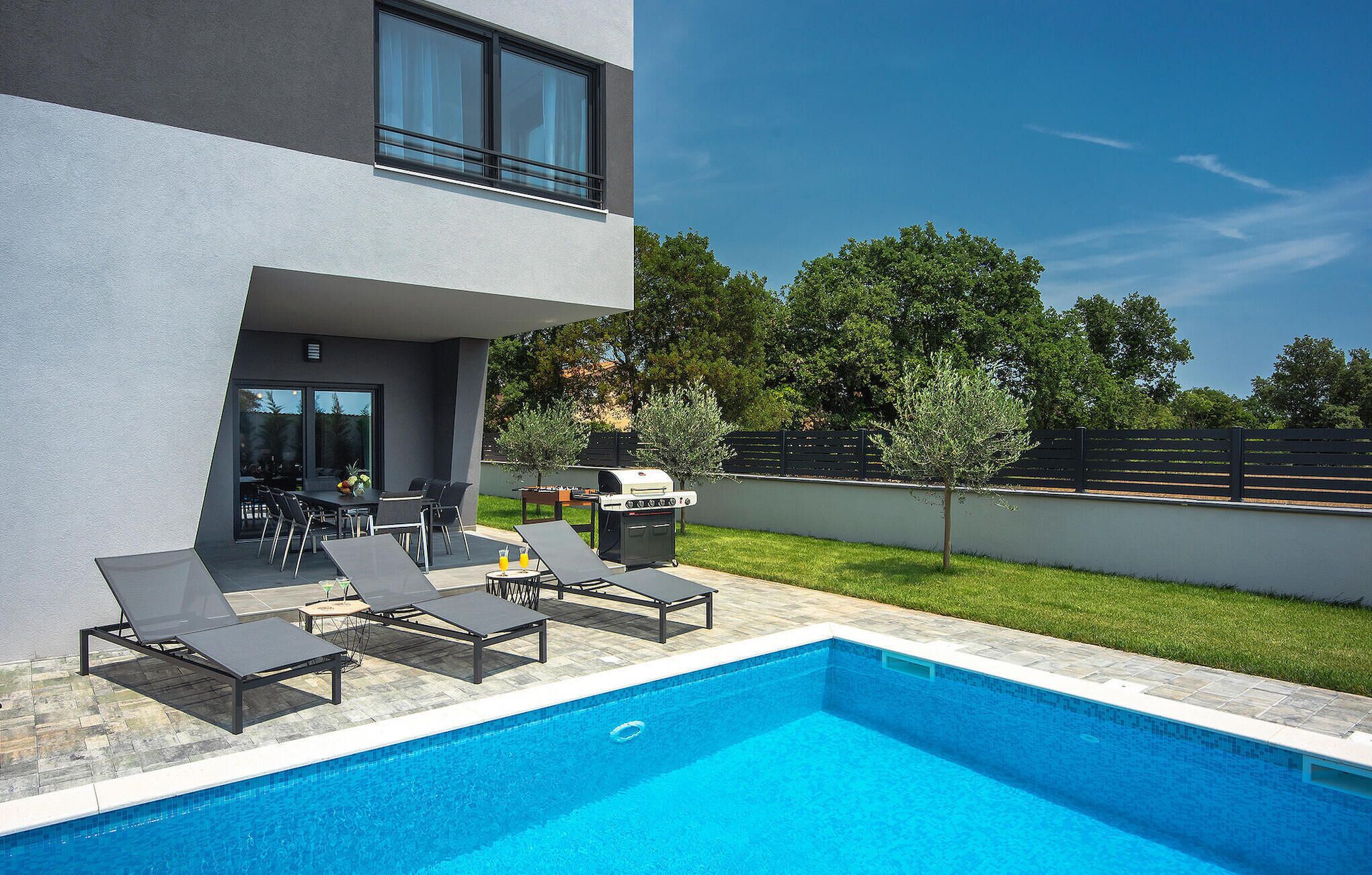 A modern outdoor pool area with lounge chairs, a barbecue grill, and a dining table.