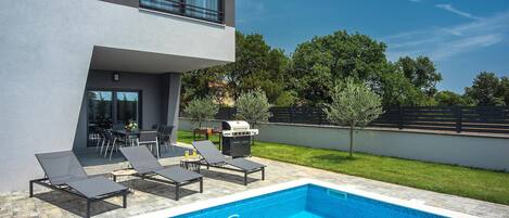 A modern outdoor pool area with lounge chairs, a barbecue grill, and a dining table.
