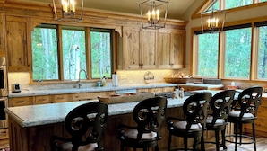 A spacious kitchen with wooden cabinets, a large island with bar stools, and large windows offering a view of the forest. - "House in the Forest" Secluded 4BD/3BA luxury home, hot tub, privacy (Crested Butte)