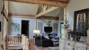 A rustic interior with exposed wooden beams, a dining area with a wooden table, a living room with a sofa and a television, and a staircase leading to an upper level. - "House in the Forest" Secluded 4BD/3BA luxury home, hot tub, privacy (Crested Butte)