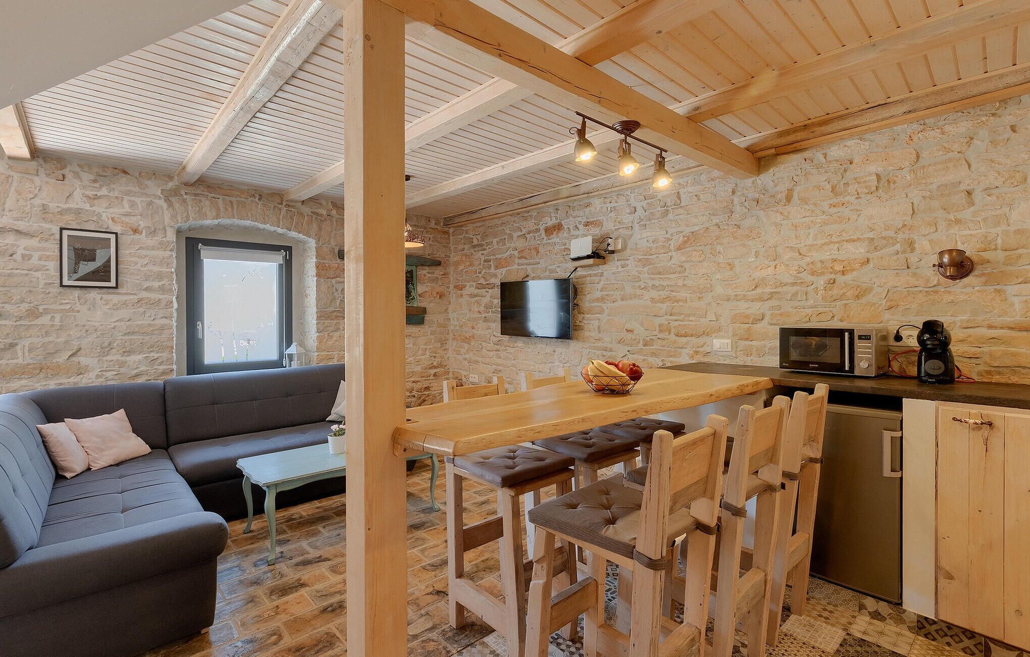 Une pièce chaleureuse avec un mur en pierre, un plafond en bois, un canapé, une table à manger avec des chaises, un téléviseur, un four à micro-ondes et une machine à café.