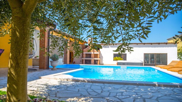 A swimming pool with stone tiles, surrounded by a garden and a building with a tiled roof.