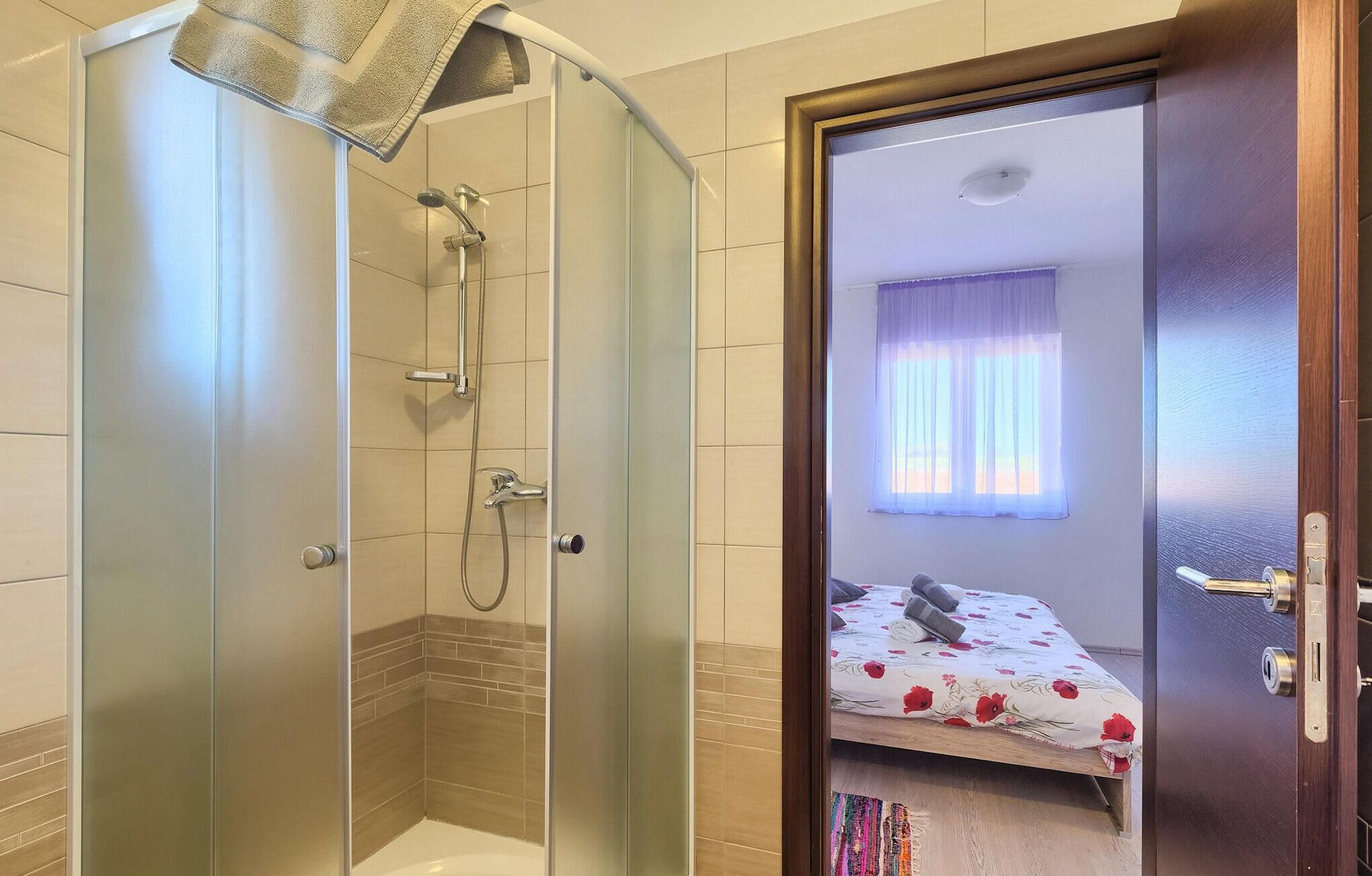 A bathroom with a glass-enclosed shower, a visible toilet, and a door leading to a bedroom with a bed and a window.