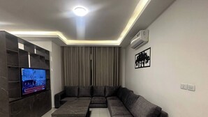 A modern living room with a sectional sofa, a flat-screen TV, a marble-tiled floor, and recessed ceiling lighting. - JRE Cozy Home Near All Services 19 (Amman)
