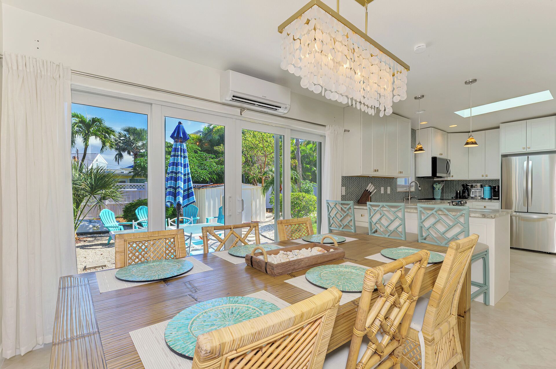 A spacious kitchen with modern appliances, a dining area with wicker chairs and a wooden table, and large sliding glass doors leading to a tropical outdoor area.
