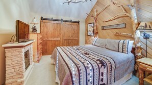 A cozy cabin-style bedroom with a stone fireplace, a large bed with patterned bedding, a wooden ceiling, and a rustic decor theme. - Mammoth Lakes Condo - Steps to Ski Lift! (Mammoth Lakes)