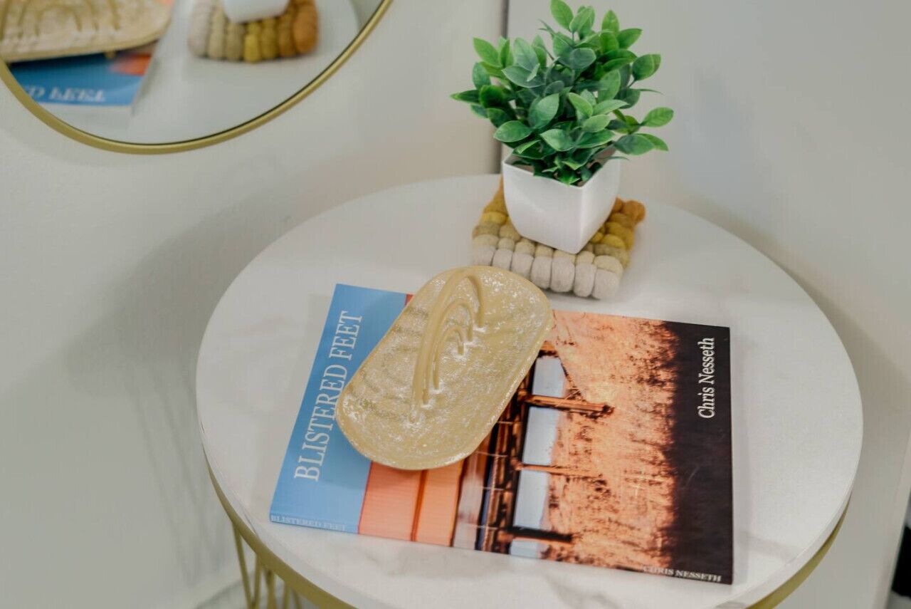 A round table with a book titled 'Blistered Feet' by Chris Nesser, a small potted plant, and a pair of slippers.
