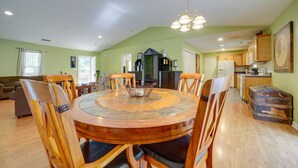 Uma ampla área de refeições com uma mesa redonda de madeira rodeada por cadeiras, um lustre e uma cozinha ao fundo.