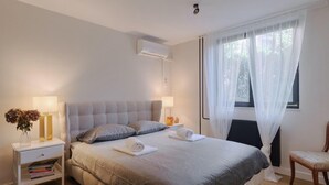 Une chambre à coucher bien rangée, avec un lit, des tables de chevet, une chaise et une fenêtre ornée de rideaux légers.