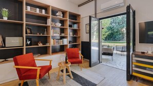 Un salon moderne avec une grande bibliothèque, deux fauteuils rouges, une petite table avec un verre à vin, et une vue sur l’extérieur grâce à une porte vitrée.