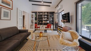 Un salon moderne avec un canapé marron, une table basse en verre, un fauteuil rouge, une grande bibliothèque et un téléviseur fixé au mur.