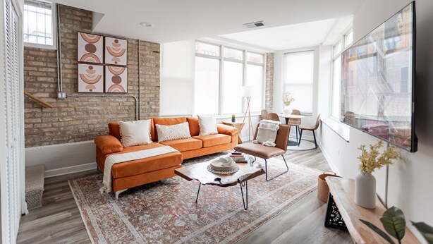 A modern living room with a brick wall, orange sofa, and a large window. - Duplex 2 Bedroom and 1.5 Bath Retreat Near Loyola Park (Chicago)