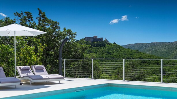 A swimming pool with lounge chairs and an umbrella, surrounded by a metal fence, with a view of a castle on a hill. - Cozy home in Drivenik (Drivenik)