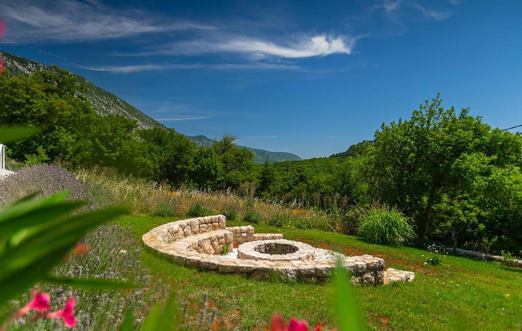 A stone ruin with a circular structure in a grassy field with trees and mountains in the background.