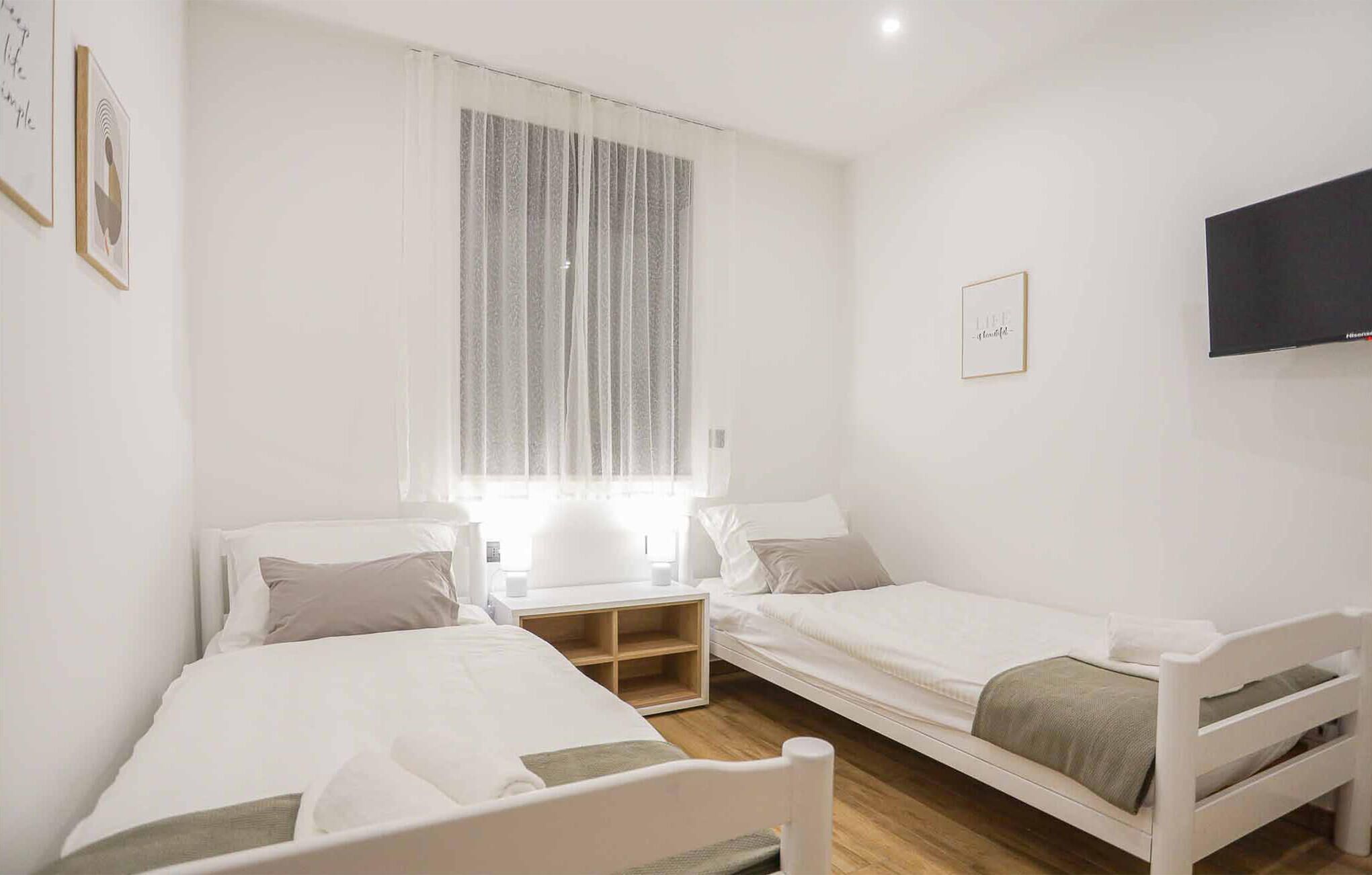 A bedroom with two single beds, a bedside table, a TV mounted on the wall, and a window with sheer curtains.