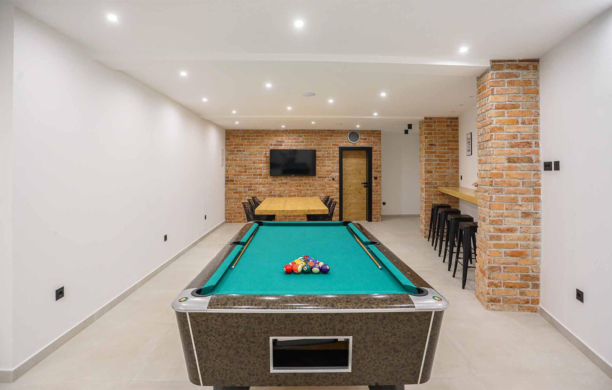 A pool table with balls in a room with brick walls, a TV mounted on the wall, and a door leading to another room.