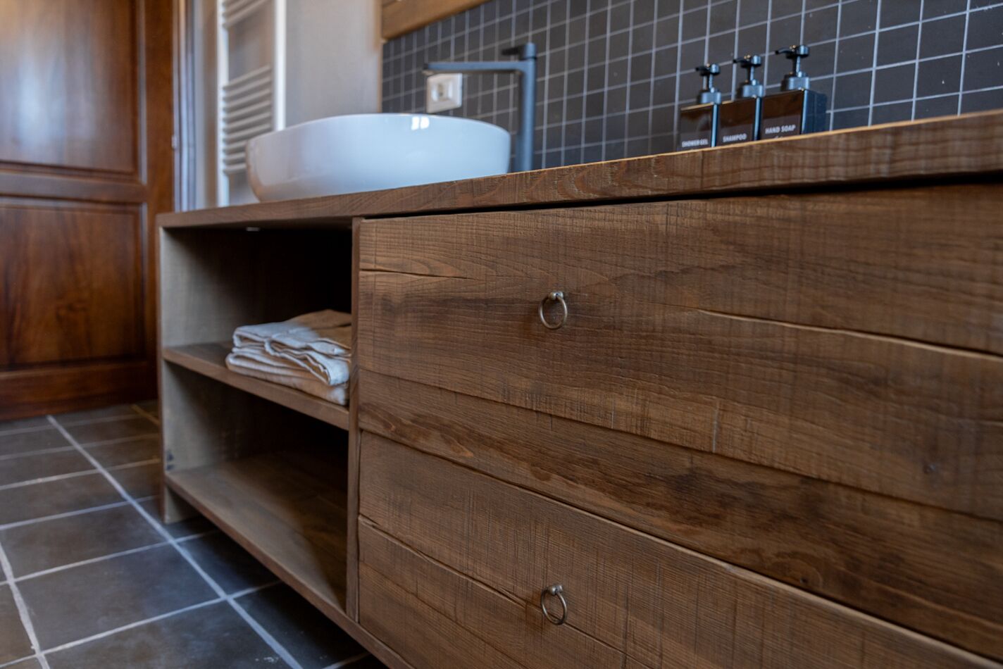 A wooden vanity with a sink, a shelf with folded towels, and a tiled floor.