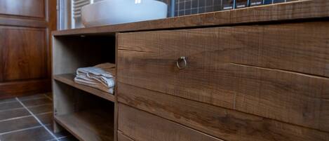 A wooden vanity with a sink, a shelf with folded towels, and a tiled floor.