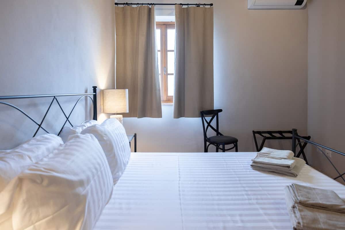 Habitación de hotel con cama, una silla, una mesa, una lámpara y ventana con cortinas.