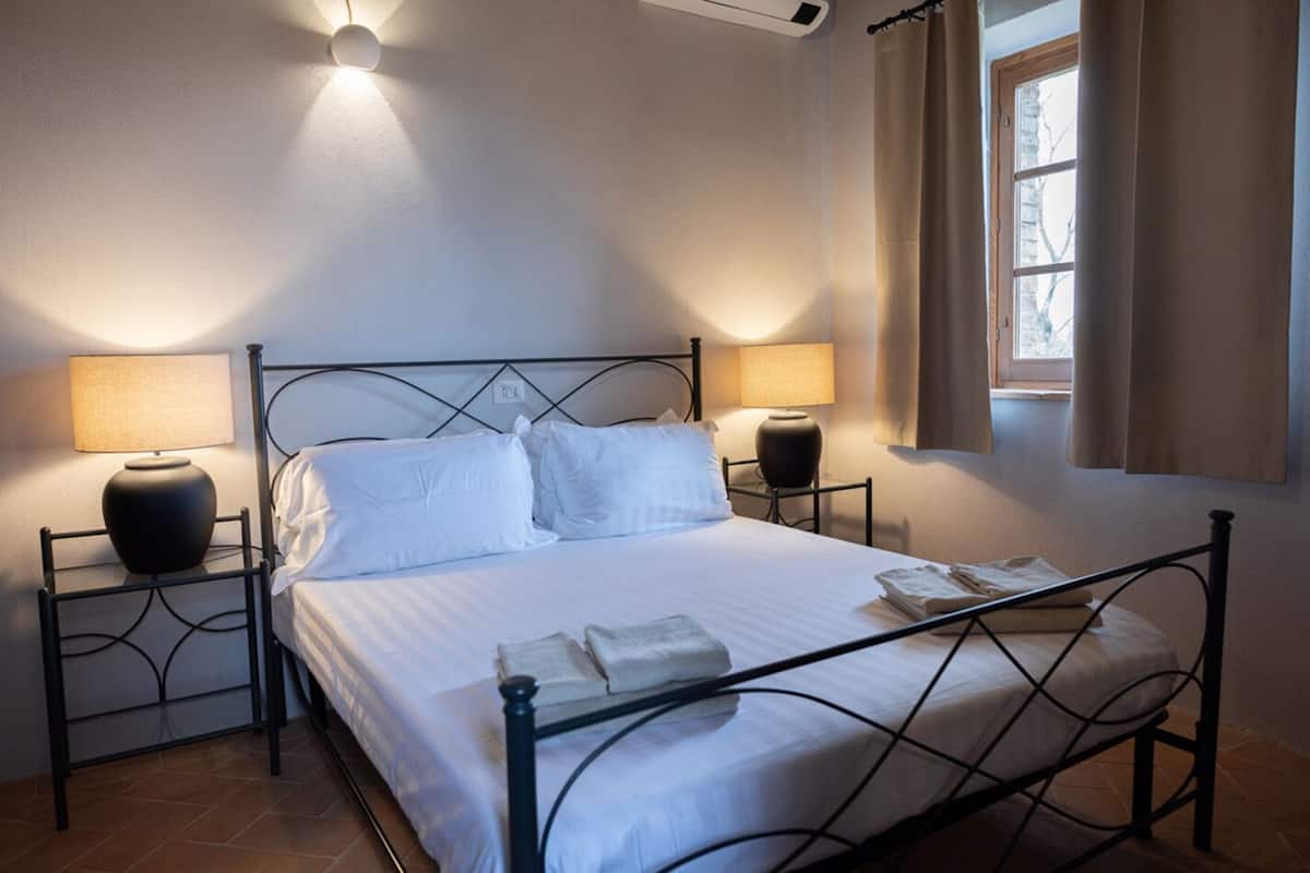 Una cama bien hecha con sábanas blancas, dos mesitas de noche con lámparas y una ventana con cortinas.