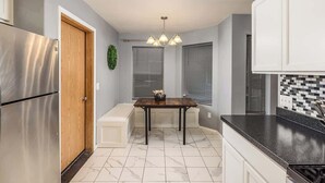 Eine moderne Küche mit einem Edelstahl-Kühlschrank, einer Holztür, einem Esstisch mit Stühlen, einer Pendelleuchte und einer Fliesenküchenwand.