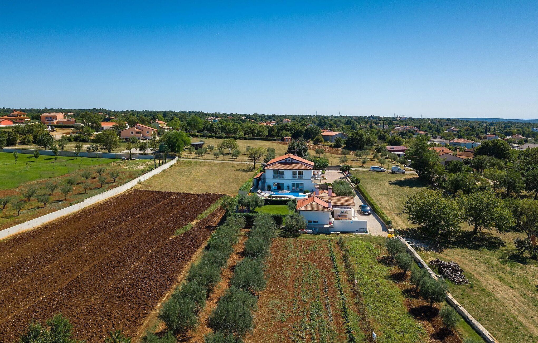 A large two-story house with a red-tiled roof, surrounded by a well-maintained garden and fields.