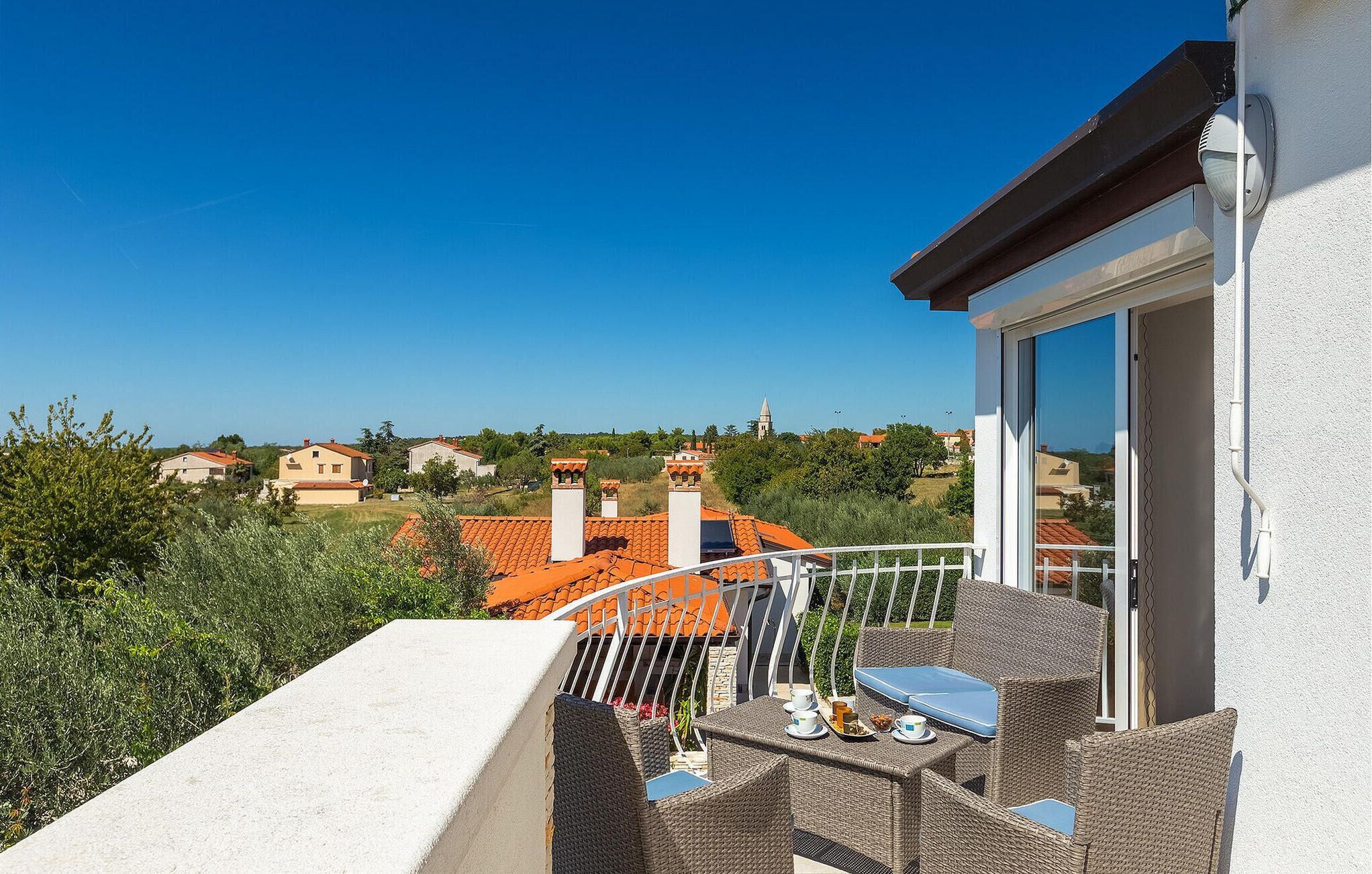 A balcony with wicker furniture and a table, overlooking a residential area with red-tiled roofs.
