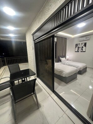 A modern room with a glass door leading to a balcony, a bed with two pillows, a small table with chairs, and a wall-mounted air conditioner. - JRE Cozy Home Near All Services 20 (Amman)