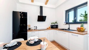 A modern kitchen with white cabinets, a black refrigerator, a microwave, and a sink with a black faucet. There is a wooden dining table with a black placemat, and a window with a view of trees.