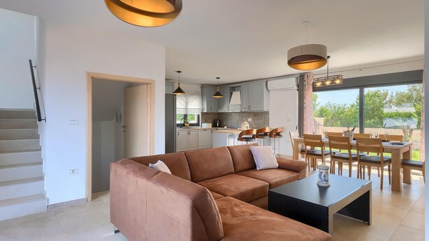 Ein modernes Wohnzimmer mit einer braunen Ledersofa, einem schwarzen Couchtisch und einem Essbereich mit Holzsesseln und Tisch. Über der Couch und dem Esstisch befinden sich Pendelleuchten.