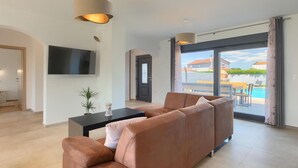 Ein modernes Wohnzimmer mit Sofa, Fernseher, kleinem Tisch mit Pflanze und Blick auf Pool und Häuser durch große Fenster.