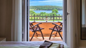 Una habitación con cama, ventana con vista a un lago y vegetación, y dos sillas afuera.