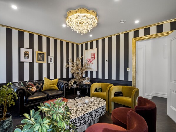 A room with a striped wall, a black leather sofa with yellow cushions, a round coffee table, and a chandelier.