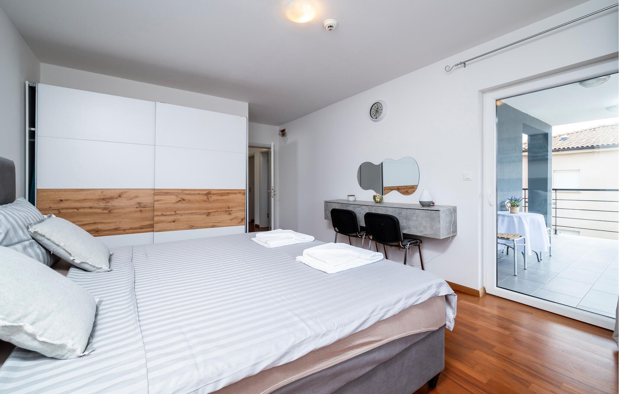 Een moderne slaapkamer met een groot bed, een bureau met stoelen, een spiegel en een schuifdeur naar een balkon.