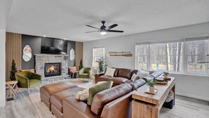 A modern living room with a stone fireplace, leather sofas, and large windows. - Poconos Luxury Retreat - Hot-Tub, Billiards, Game Room, Fire-Pit, Private! (Tobyhanna)