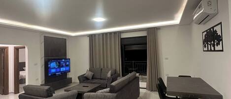 A modern living room with a flat-screen TV, grey sofas, a dining table with chairs, and an air conditioning unit on the wall.