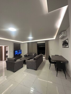 A modern living room with a flat-screen TV, grey sofas, a dining table with chairs, and an air conditioning unit on the wall. - JRE Cozy Home Near All Services 21 (Amman)