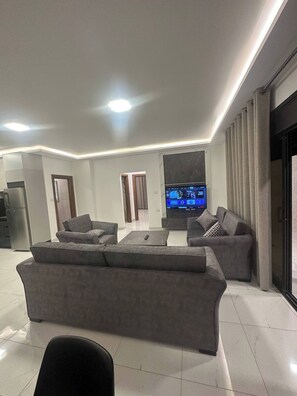 A modern living room with a large flat-screen TV, grey sofas, a coffee table, and a kitchen area with a refrigerator. - JRE Cozy Home Near All Services 21 (Amman)