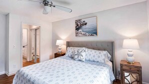 A bedroom with a bed, two bedside tables with lamps, a ceiling fan, a framed picture on the wall, and a door leading to another room. - Ocean Walk Condo Upstairs Unit (St. Simons Island)