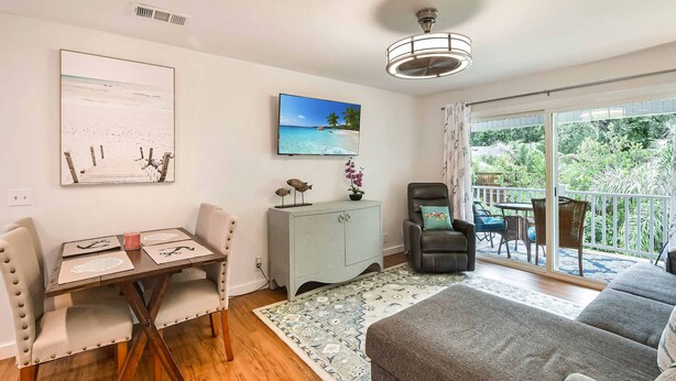 A modern living room with a dining area, a grey sofa, a dining table with chairs, a grey cabinet, and a TV on the wall. - Ocean Walk Condo Upstairs Unit (St. Simons Island)