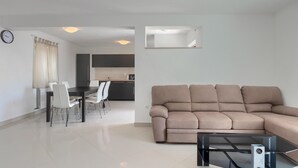 Una sala moderna con un sofá beige, un comedor con sillas blancas y una cocina al fondo.