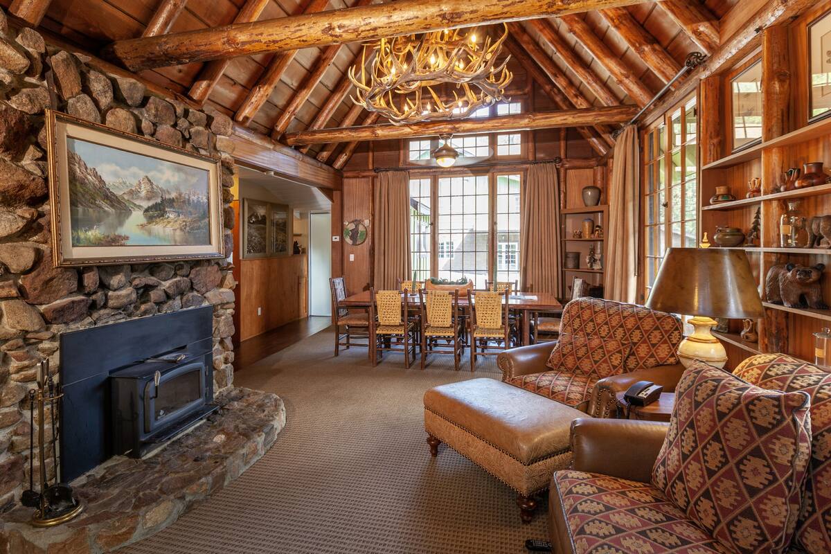 A rustic interior with a stone fireplace, wooden ceiling beams, a chandelier, and a dining area with a table and chairs.