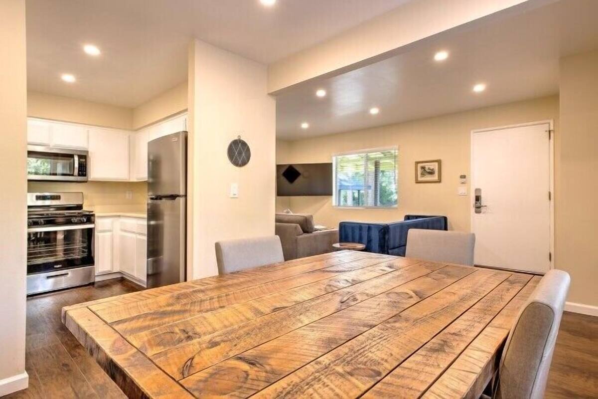 A modern kitchen with stainless steel appliances, a dining area with a wooden table, and a living space with a sofa and a window.