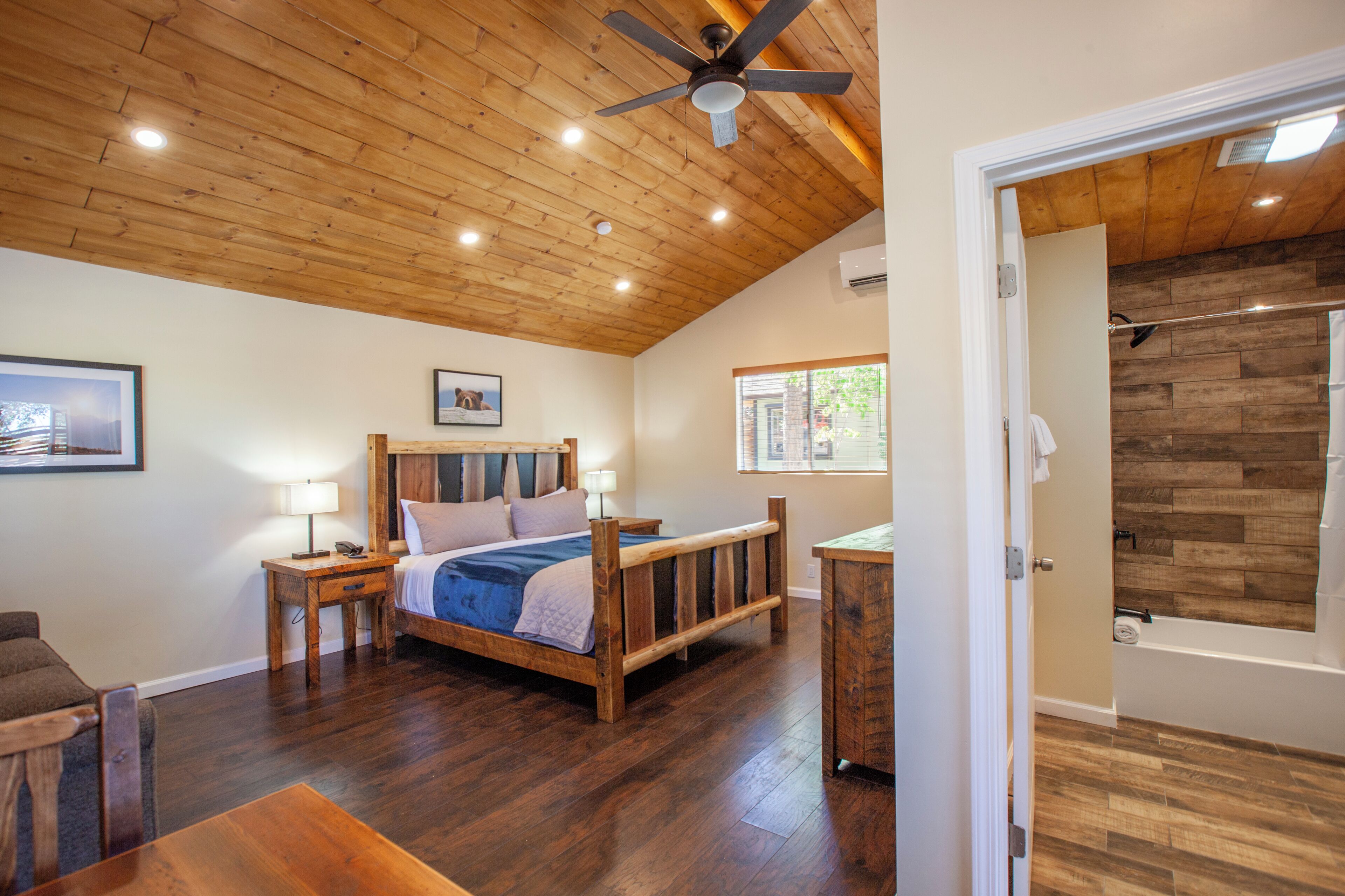 Un dormitorio con techo de madera, una cama con cabecera, mesitas de noche, suelo de madera y vistas a un baño con pared de madera.