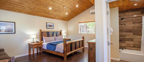Un dormitorio con techo de madera, una cama con cabecera, mesitas de noche, suelo de madera y vistas a un baño con pared de madera.
