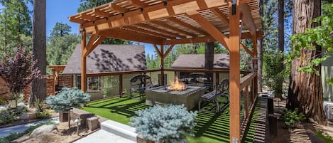 A wooden pergola with a fire pit and seating area, surrounded by a landscaped garden with trees and a lawn.